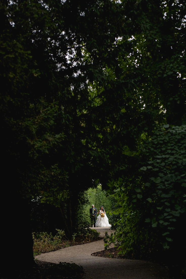 A bride walks down the aisle on the arm of her father in the Outdoor Ceremony Garden surrounded by trees. 